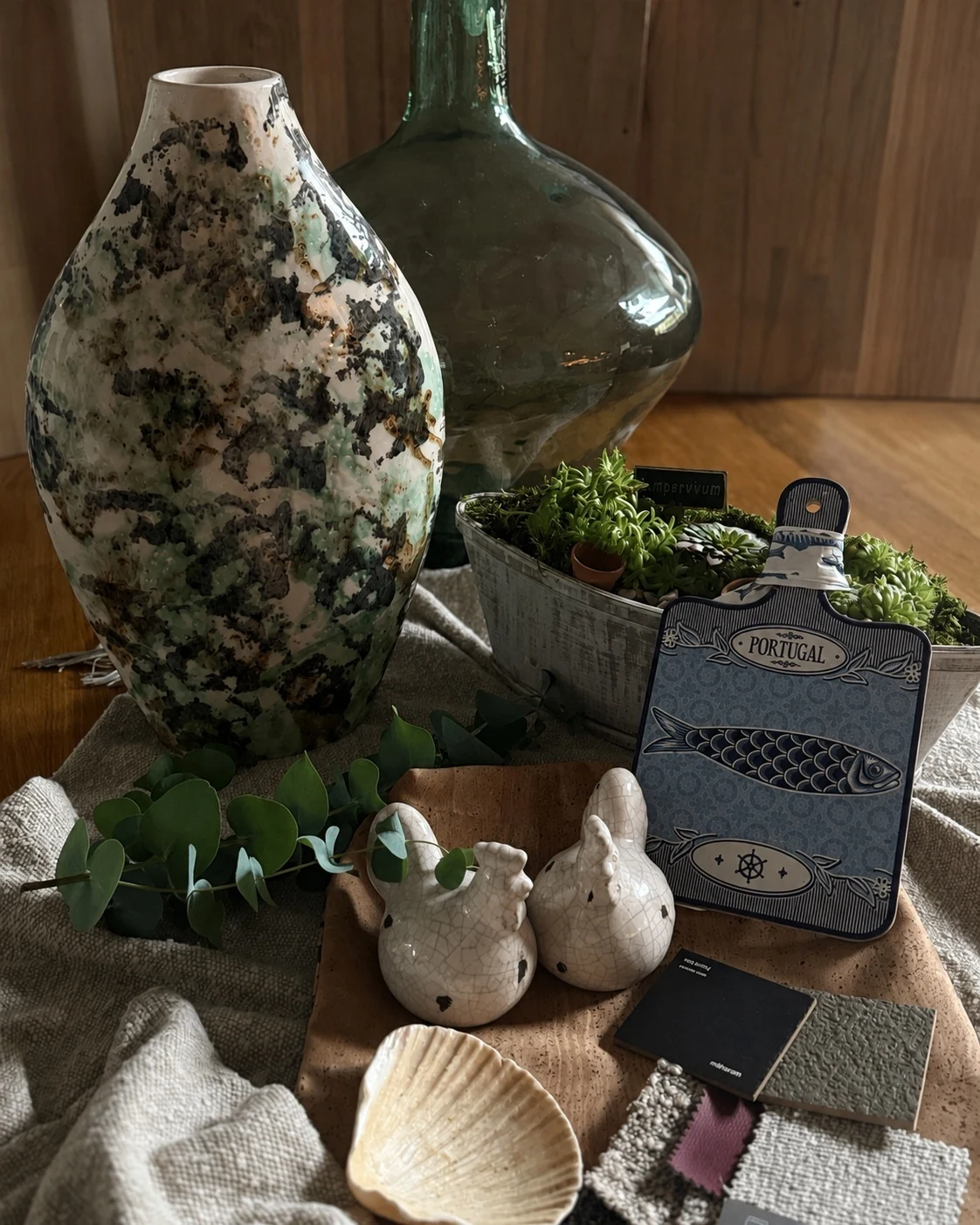 A composition of Portuguese materials and objects: a hand-glazed ceramic vase, a green glass demijohn, cork, linen, wool upholstery samples, small ceramic birds and a blue decorative board with a sardine motif, arranged in natural shades of green, beige and wood.