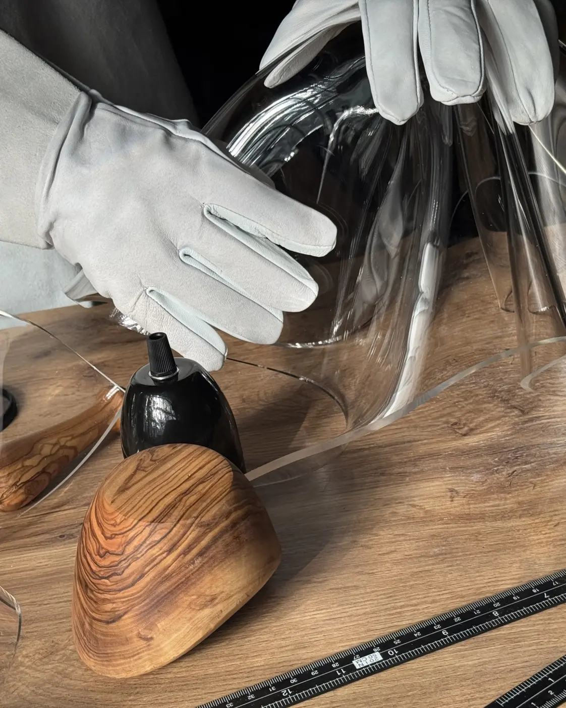 Hände in hellen Lederschutzhandschuhen legen eine transparente Acrylglasplatte auf eine hölzerne Werkbank; das Material fällt in weichen Falten. Darunter: eine schwarze Lampenfassung, glatte Holzformen und ein Stahlwinkel.